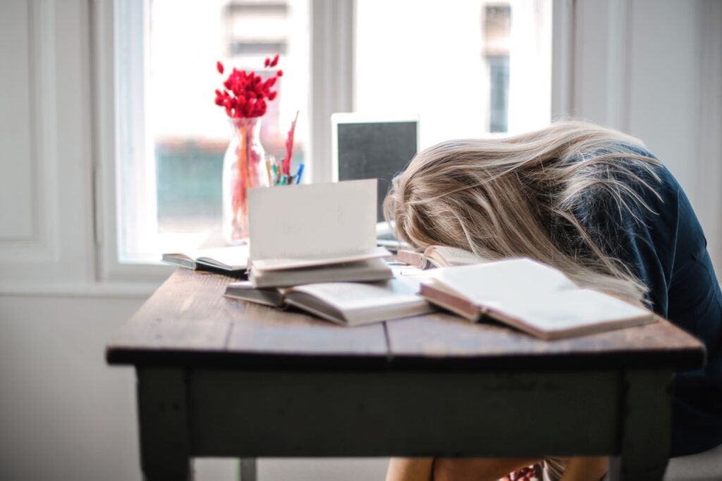 Blonde woman slumped over desk filled with books, showcasing procrastination and study fatigue