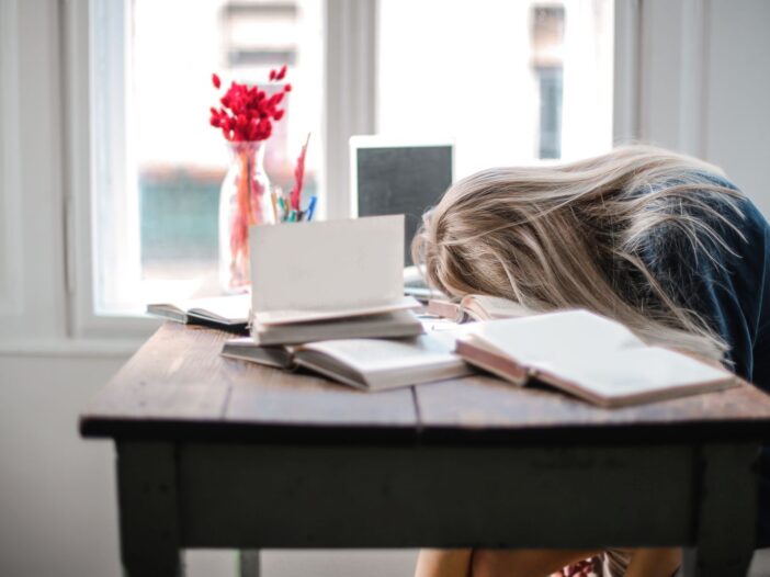 Blonde woman slumped over desk filled with books, showcasing procrastination and study fatigue