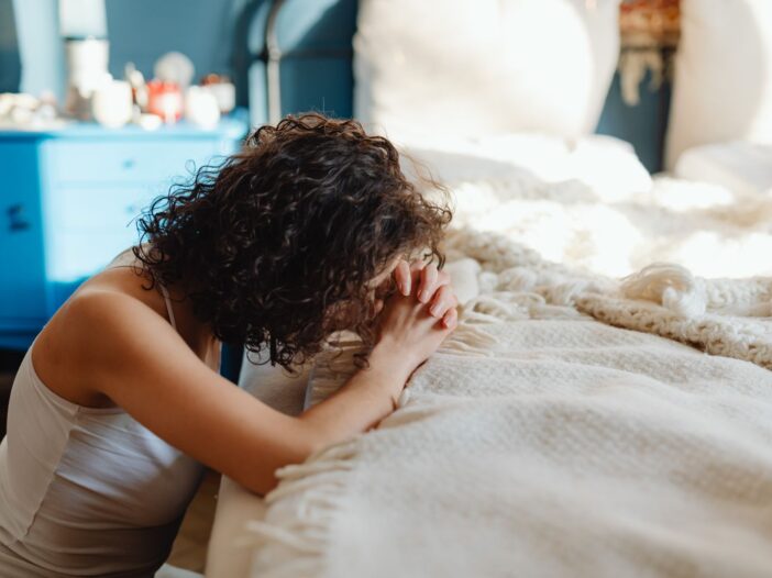 A woman kneels in contemplation by her bed, showcasing spirituality and tranquility in a warmly lit bedroom.