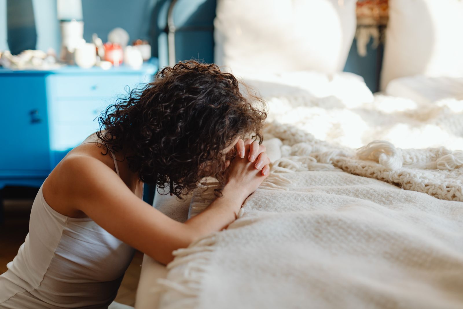 A woman kneels in contemplation by her bed, showcasing spirituality and tranquility in a warmly lit bedroom.