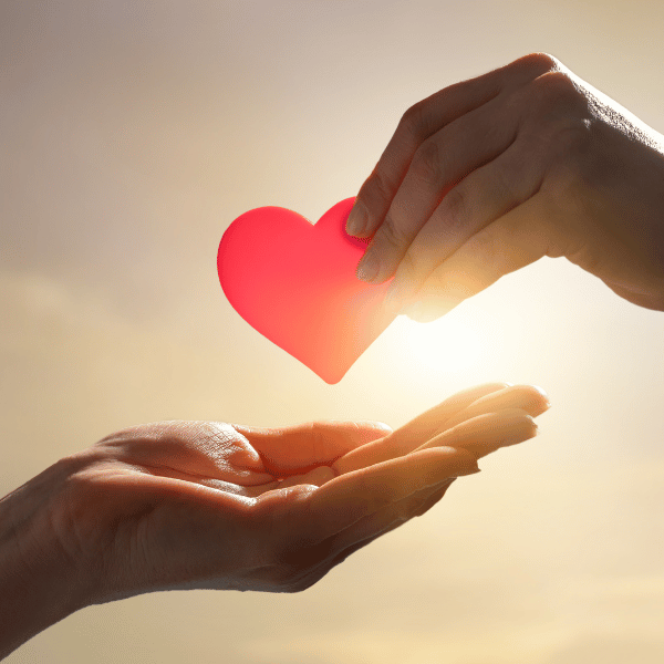 Two hands with one handing a heart to another against a sunny background