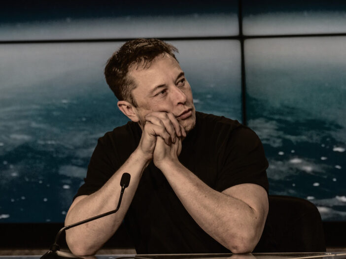 Interview setting for Elon Musk AI article, showing a man seated at a table with an Earth backdrop.