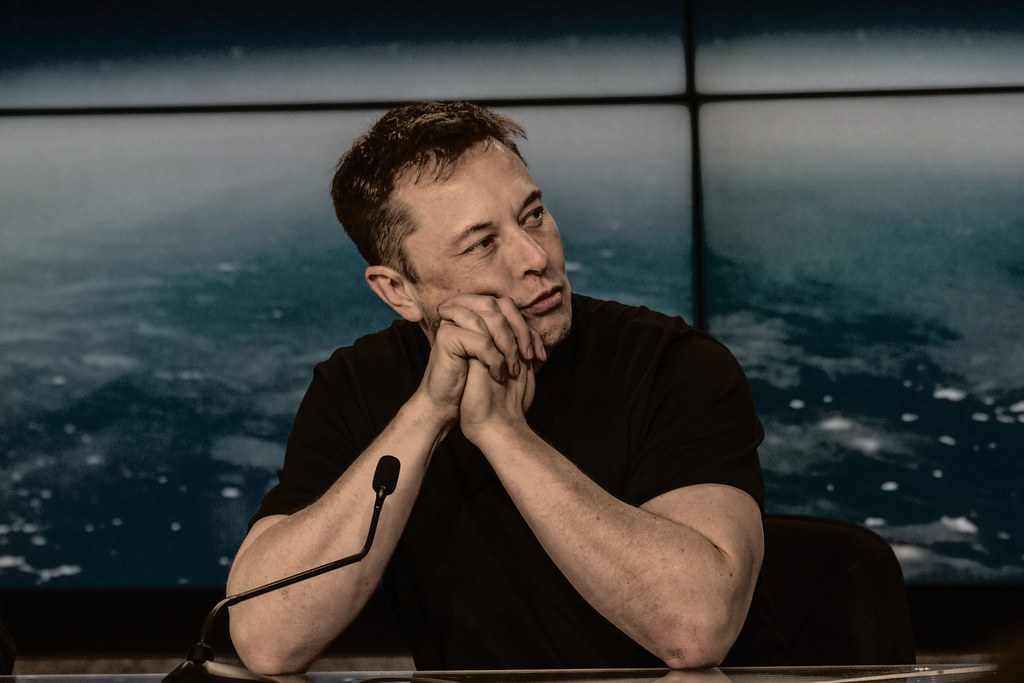Interview setting for Elon Musk AI article, showing a man seated at a table with an Earth backdrop.