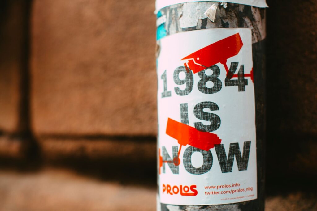 Street poster reading “1984 Is Now” with red surveillance camera symbols