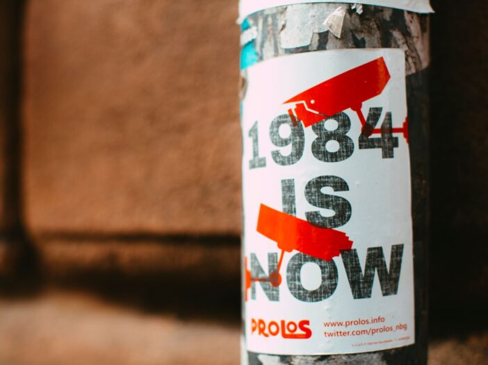 Street poster reading “1984 Is Now” with red surveillance camera symbols