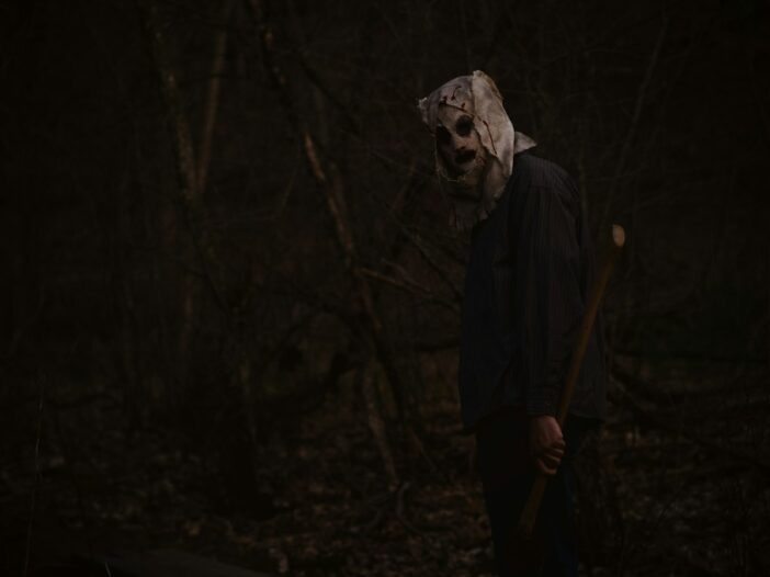 Masked horror figure standing in a dark forest to illustrate a collection of Halloween movie quotes.