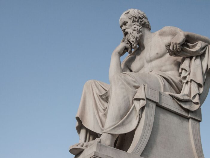 Stone statue of philosopher Socrates sitting in thoughtful pose in Athens, Greece under a clear blue sky.
