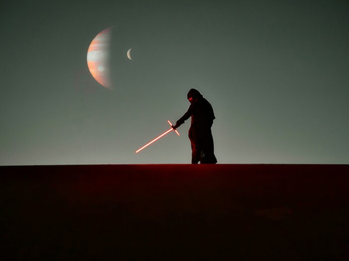 Silhouetted figure holding a red crossguard lightsaber beneath distant planets in a Star Wars-inspired landscape