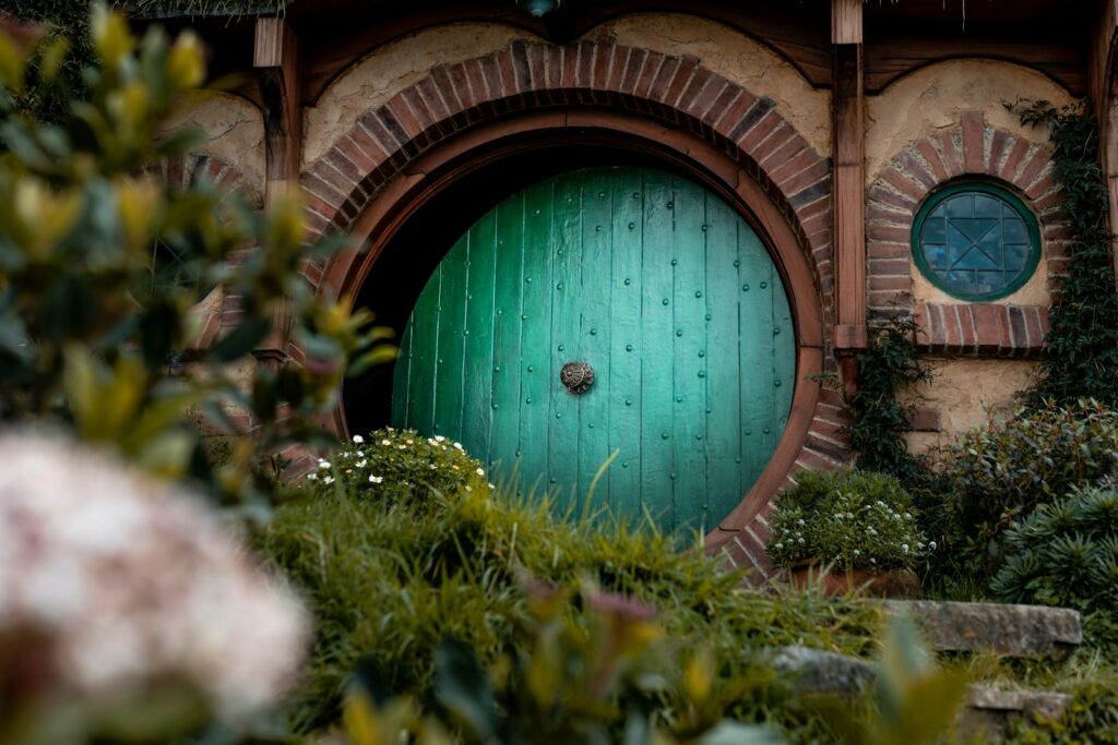 Round green door of a hobbit-style home framed by brickwork, flowers, and lush greenery.