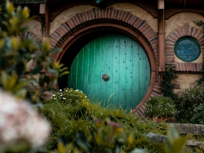 Round green door of a hobbit-style home framed by brickwork, flowers, and lush greenery.