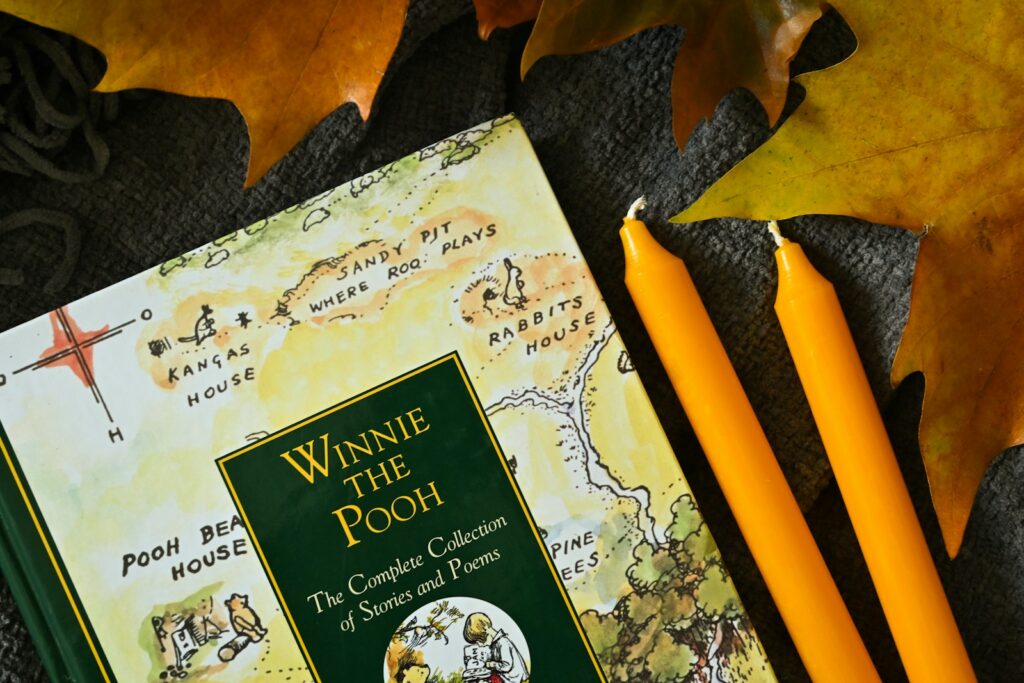 Winnie the Pooh book beside orange pencils on a dark surface with autumn leaves.