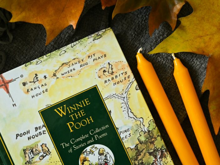 Winnie the Pooh book beside orange pencils on a dark surface with autumn leaves.