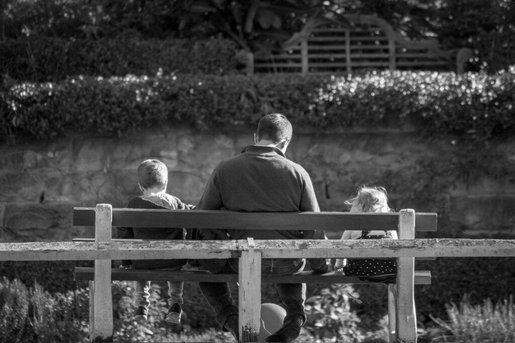 grayscale photography of father and two children sitting on a bench