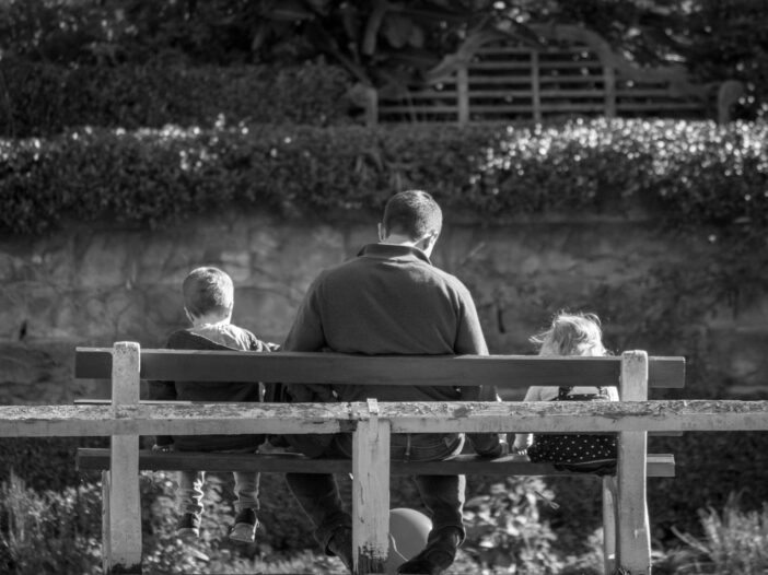 grayscale photography of father and two children sitting on a bench